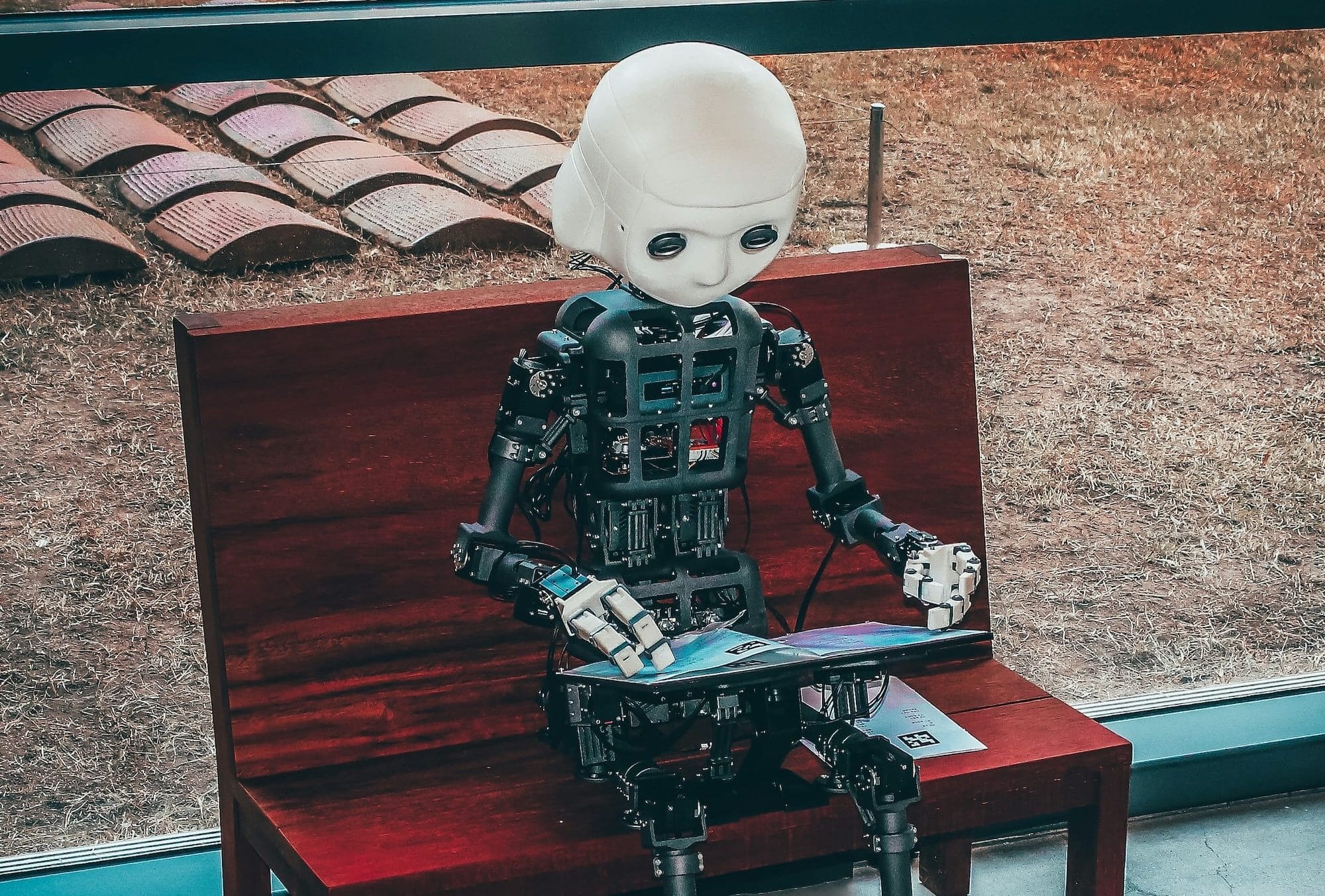 A humanoid robot with a white head and exposed mechanical parts sits on a wooden bench, holding and looking at an open magazine indoors near a window.
