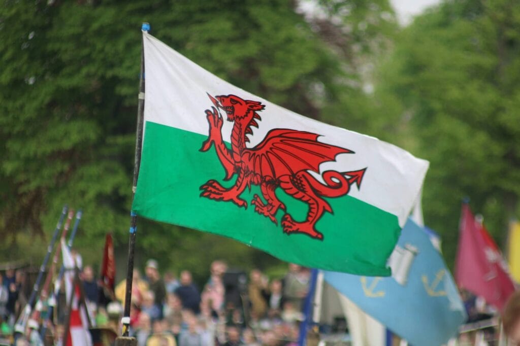 The Welsh national flag with a red dragon on a green and white background is flying outdoors, with blurred people and trees in the background.